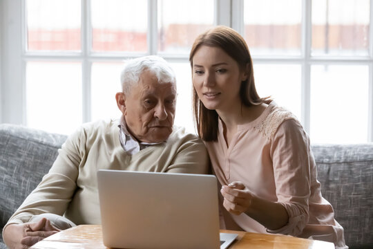 Young female social worker volunteer help assist retired elderly man in shopping online using home computer. Focused old father look at laptop screen watch grown daughter teaching him pay bills on pc