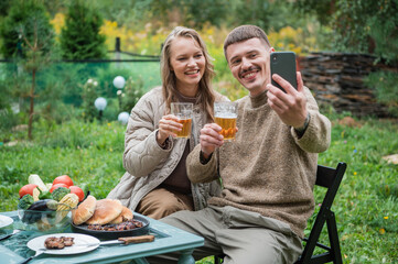 Video conference with friends in the open air during a picnic in nature with beer and barbecue. Young people use their smartphone to chat with friends and have fun