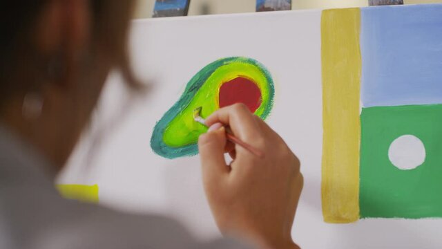 Close-up of white canvas at easel with abstract painting consisting of geometric shapes and avocado painted by unrecognizable woman