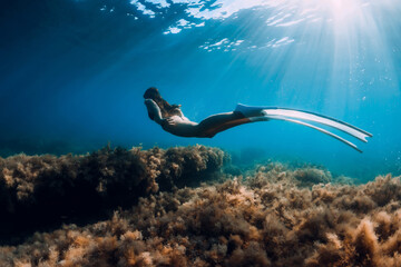 Freediver girl with white fins glides underwater with amazing sun rays and seaweed. Free diving underwater in blue sea