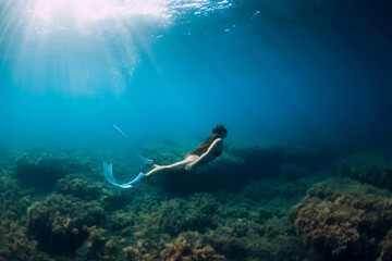 Freediver girl with white fins glides underwater with amazing sun rays and seaweed. Free diving underwater in blue sea