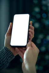 Smartphone mockup template with white screen and bokeh effect background. A young woman uses her smartphone to socialize, wishes and celebrate with friends online from her living room.