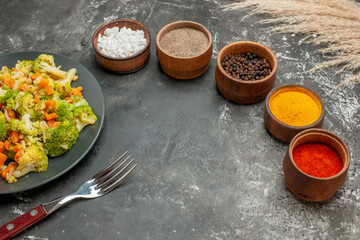 Top view of set of different spices in brown bowls and vegetable salad on gray background