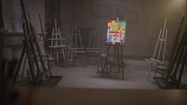 Full shot of empty dark art studio and bright abstract painting of colorful geometric shapes on canvas standing in middle of room