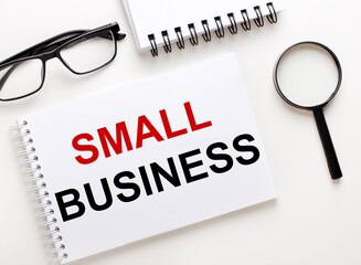 SMALL BUSINESS is written in a white notebook on a light background near the notebook, black-framed glasses and a magnifying glass.