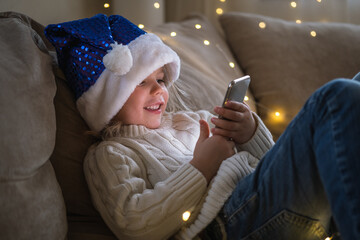 Girl in blue Santa hat using mobile phone for video call friends and grandparents.