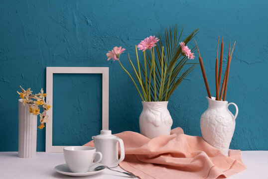 gerbera, daffodils, white frame for a photo, a cup with coffee and a teapot and two vases with a cotton plant on a blue background