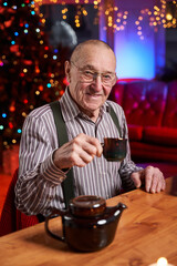 Dressed in shirt old man drink a tea sitting at table in cosy and colourful room decorated with xmas tree and lights