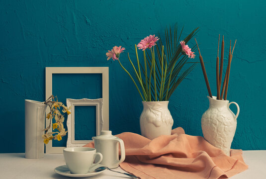 gerbera, dry daffodils, white photo frame, a cup with coffee and a teapot and two vases with flowers on a blue background