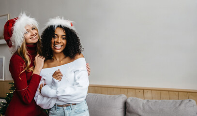Fototapeta premium Christmas unites racial diversity. African and caucasian young women with Santa hat at home hugs and looking at camera. Black lives matter and tolerance concept