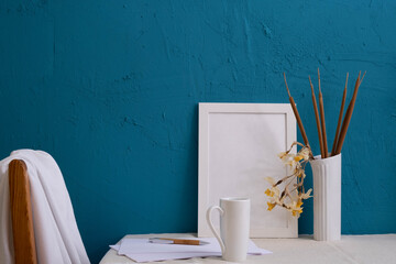 pen and a piece of paper, a vase with cones of reeds and dry daffodils, a white frame for a photo, a cup with coffee on a blue background