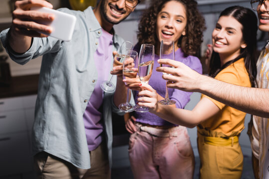 smiling hispanic man taking selfie on mobile phone with cheerful friends clinking champagne glasses, blurred foreground