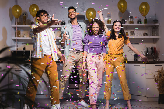 Excited Hispanic Friends Dancing In Kitchen Under Falling Confetti, Blurred Foreground