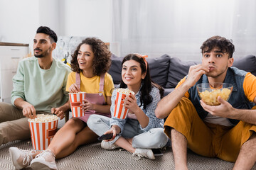 excited hispanic friends eating snacks and watching tv while sitting on floor