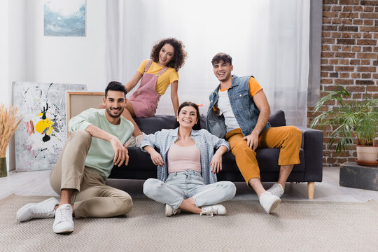 happy hispanic friends looking at camera while sitting on sofa and floor in living room