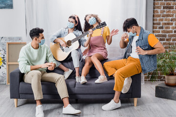 Fototapeta premium Hispanic friends in medical masks playing acoustic guitar on sofa in living room