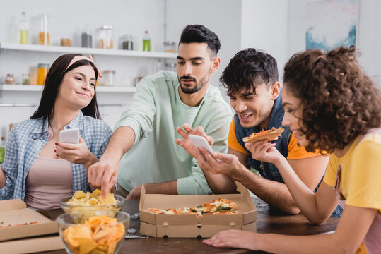 Positive hispanic friends with smartphones holding pizza and potato chips on blurred foreground at home