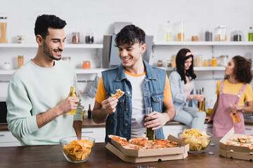Smiling hispanic friends holding beer near pizza and potato chips in kitchen