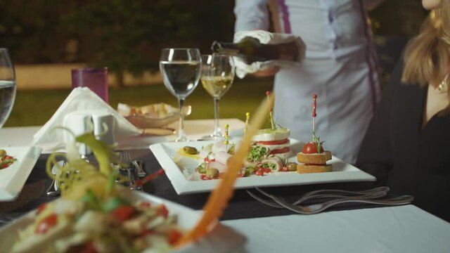 Waiter pours white wine into woman's glass at elegant dining table.