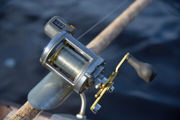 Round silver fishing reel with a line counter indicating almost 10 meters fishing line out place in a rod holder with blurred river water background.