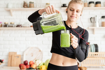 Young blond smiling woman making green smoothie at home kitchen