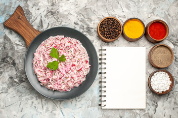 Top view of tasty chicken salad with mayonnaise and beet served with green on brown wooden cutting board and different spices lined up in a row next to notebook on ice table