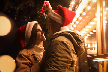 Obraz premium Happy couple in Santa hats standing under mistletoe bunch outdoors, bokeh effect