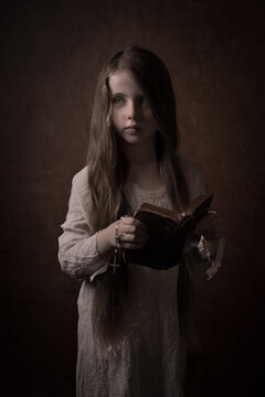 Young girl in white dress holding old antique bible and rosary in dark studio setting