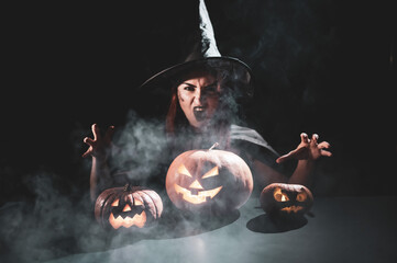 Fototapeta na wymiar The evil witch casts a spell on pumpkins. Portrait of a woman in a carnival halloween costume in the dark