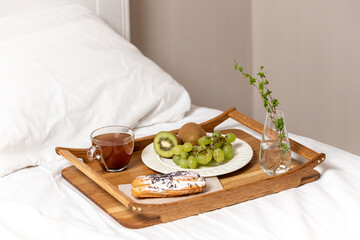 Wooden tray with a cup of tea or coffee, eclair and fruit. Breakfast in bed. Kiwi and green grapes in the morning. The concept of a good and cheerful spring morning.