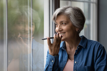 Happy middle aged elderly woman recording audio message or using voicemail mobile function, standing near window. Smiling 60s old lady activating virtual assistance or web surfing information online.