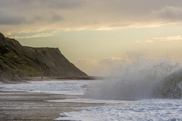 Beach scene with sandy cliffs and a big crashing wave. Few people along the beach