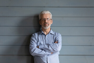 Portrait of serious pensive middle aged 60s grey haired man in eyeglasses standing with folded arms near wall. Thoughtful elderly senior confident grandfather posing for photo, retirement lifestyle.