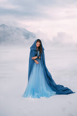 Art photo. Fantasy young woman fairy elf in blue cape with hood stands in cold wind. Winter nature background, mountains in the clouds, dramatic sky white snow. Girl Queen walks in dress, silk cloak © kharchenkoirina