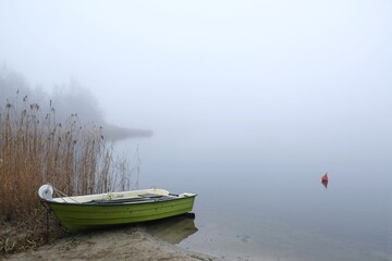 Green wooden boat on shore of lake in calm misty day. Red buoy on water.