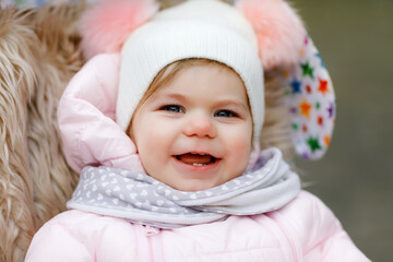 Cute little beautiful baby girl sitting in the pram or stroller on cold autumn, winter or spring day. Happy smiling child in warm clothes, fashion stylish baby coat and hat. Snow falling down