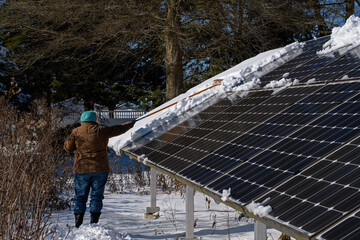 Woman cleaning snow covered solar panels on a bright and cold winter’s day.   Panels are surrounded by the remnants of a dormant perennial pollinator garden.