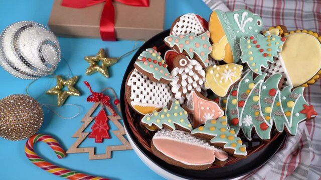 A dish of Christmas gingerbread with different shapes-snowflakes, firs, mittens, stars, sleigh, cones, bells rotates on the table, blue background. Festive decorations all around.