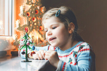 Litte toddler girl sitting by window and decorating small glass Christmas tree with tiny xmas toys. Happy healthy child celebrate family traditional holiday. Adorable baby.