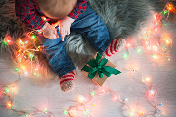 Baby boy in a plaid shirt sitting near a garland holding a box with a gift