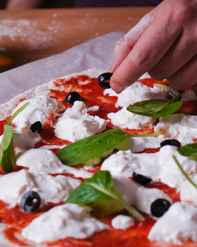 Vertical shot of a cook putting an olive on the Artisan pizza dough with mozar