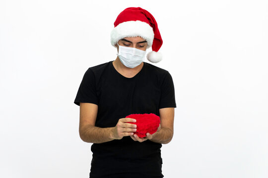 Young male wearing a medical face mask and a santa hat. Young man is holding a heart-shaped gift box in his hands. Isolated image and white background.