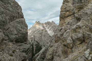 Fototapeta premium A sneak-peak on a high and distant mountain in Italian Dolomites. The mountain is visible between two rocky and steep slopes in front. Playing hide and seek. Raw and desolate landscape. Overcast