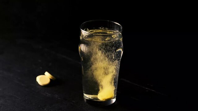 An effervescent orange vitamin C tablets falls into a glass of water and dissolves by stirring the water with its bubbles on the black background. Stay healthy and fight agains covid virus.