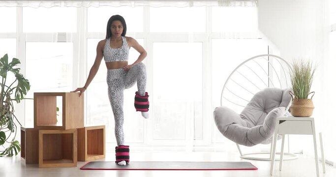 Pretty fitness black woman in sportswear practicing donkey kick exercise with ankle weight for glutes strength training at home during covid-19.