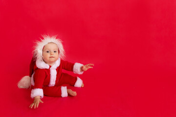 a small child of six months in a Santa costume sits on a red isolated photophone, a place for text, the concept of the new year and holiday