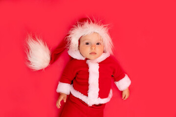 a small child of six months in a Santa costume lies on a red isolated photophone, a place for text, the concept of the new year and holiday