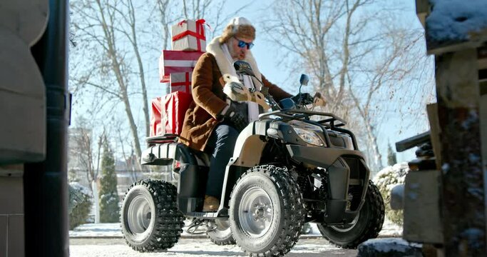 Modern Santa Claus. Cheerful Santa Claus delivers gifts on an ATV on Christmas day on the background of a snow house the background.