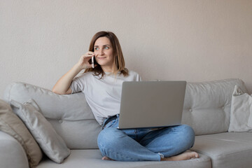 Portrait of female freelancer working at home with laptop and mobile phone. Happy woman surfing and browsing net and talking on smartphone, sitting on couch, relaxing in living room.