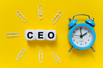 On a yellow background, a blue alarm clock, paper clips and white cubes on which the text is written - CEO